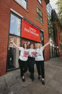 Amelia Sutcliffe, Sammi Allen and Katie Bullimore stand in front of the Smith Partnership Solicitors head office in Friar Gate Derby wearing T-shirts with sponsors names on