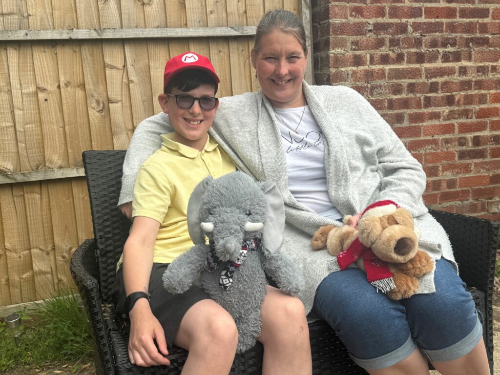 "A smiling boy wearing a red cap and yellow shirt sits next to a woman in a grey cardigan. They are seated on a wicker bench outdoors. The boy is holding a grey stuffed elephant, and the woman is holding a brown plush dog wearing a red scarf and hat."