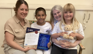Two pupils from St Chad's CofE Nursery and Infant School sit with two members of staff, proudly displaying the school's ARC Alex Timpson Attachment and Trauma Award after being named the East Midlands regional winner.