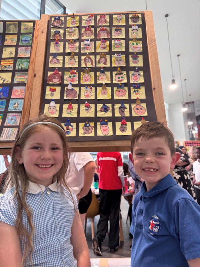 Two pupils smile beside a display of their classmates' self-portrait artwork at the Derby Diocesan Academy Trust art exhibition celebrating creativity.