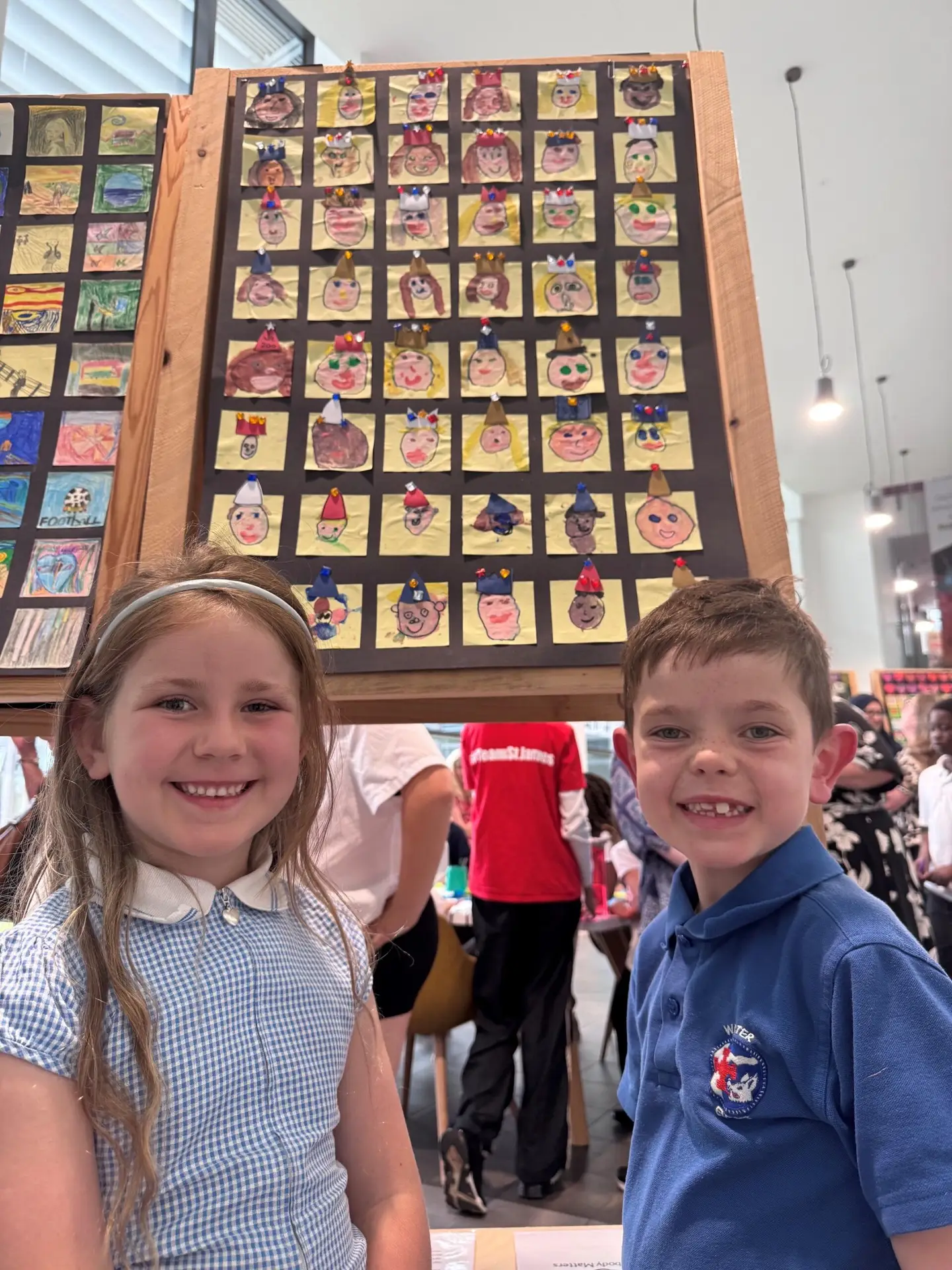 Two pupils smile beside a display of their classmates' self-portrait artwork at the Derby Diocesan Academy Trust art exhibition celebrating creativity.