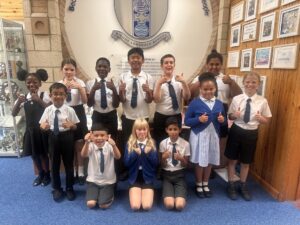A group of 13 primary school pupils in school uniform stand and kneel beneath the Bishop Lonsdale School crest, smiling and giving thumbs up in the school entrance hall.