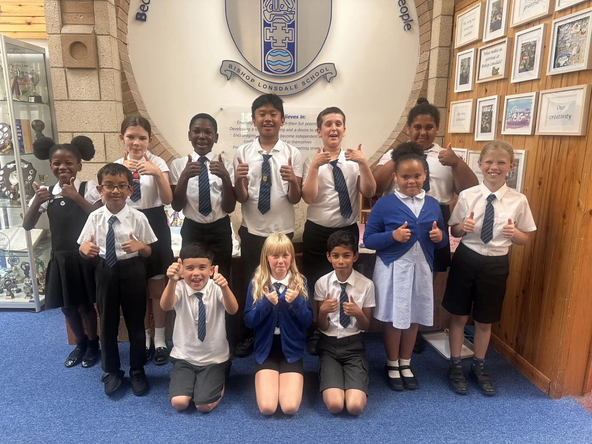 A group of 13 primary school pupils in school uniform stand and kneel beneath the Bishop Lonsdale School crest, smiling and giving thumbs up in the school entrance hall.