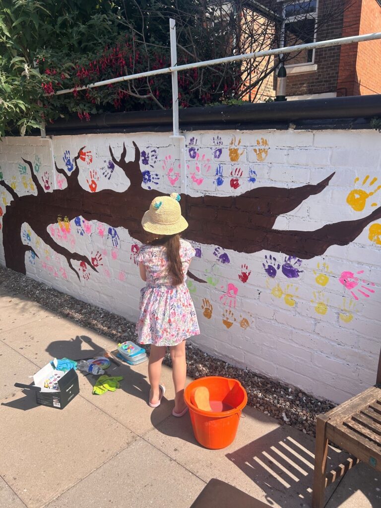 A young girl in bright clothing adds colourful handprints to a mural on a wall. The mural consists of a painted tree with handprints representing leaves.
