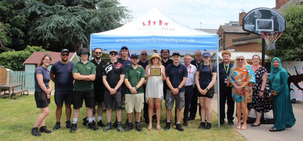 A team of apprentices stand beneath a Children First Derby gazebo holding a plaque. They are stood within a bright and colourful playground.