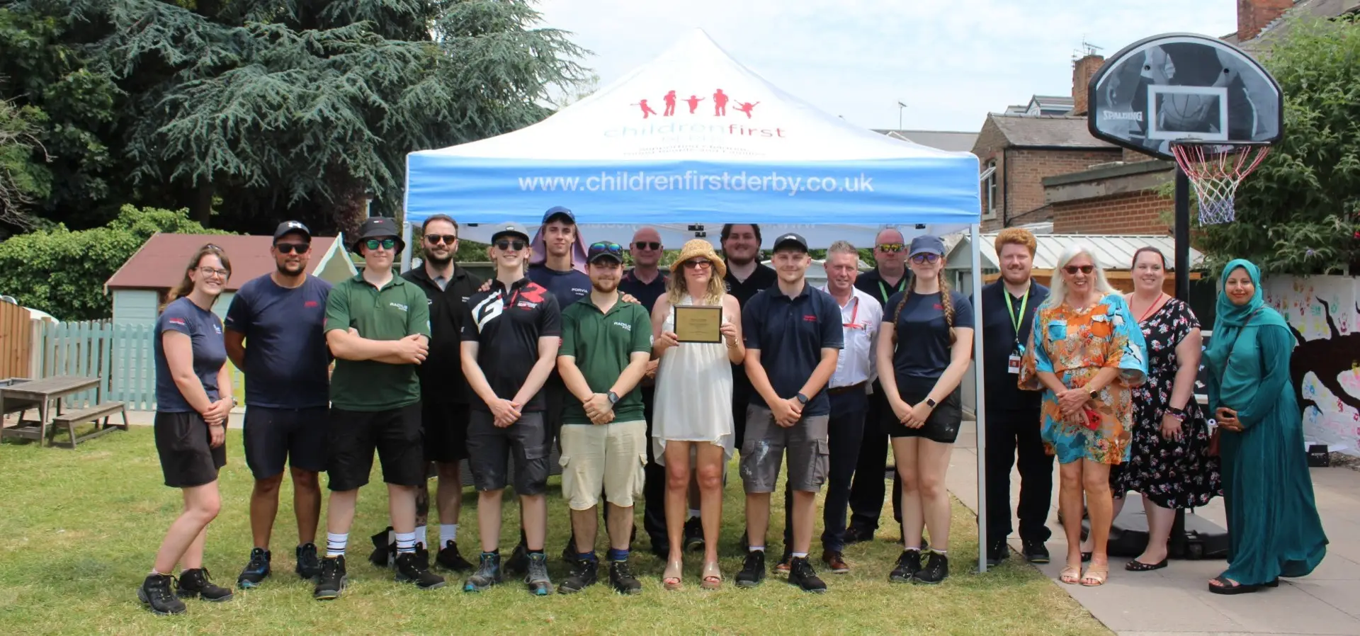 A team of apprentices stand beneath a Children First Derby gazebo holding a plaque. They are stood within a bright and colourful playground.