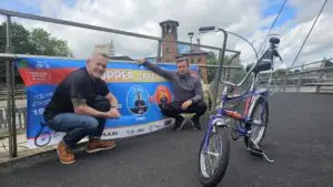 Dean Jackson MBE and Phil Mason crouch beside a promotional banner for the Chopper Canal Challenge with a purple Raleigh Chopper bicycle in the foreground. The banner advertises a 100-mile charity ride from Preston Brook in support of YMCA, while the Derby Silk Mill is visible in the background.