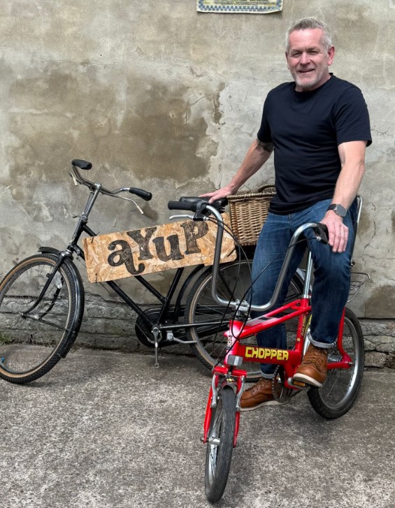 Dean Jackson MBE sits on a red vintage Raleigh Chopper bicycle beside a black utility bike with a wooden "ayup" sign attached to the frame.
