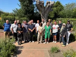 A group photo of Growing Lives participants, staff and supporters standing together in the centre's garden on a sunny day beneath a large pollarded tree.