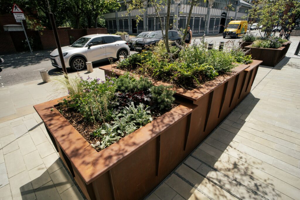 Large rectangular steel planters filled with trees and flowering plants line a wide paved sidewalk beside a city street, creating a green pedestrian space with parked cars and buildings in the background.