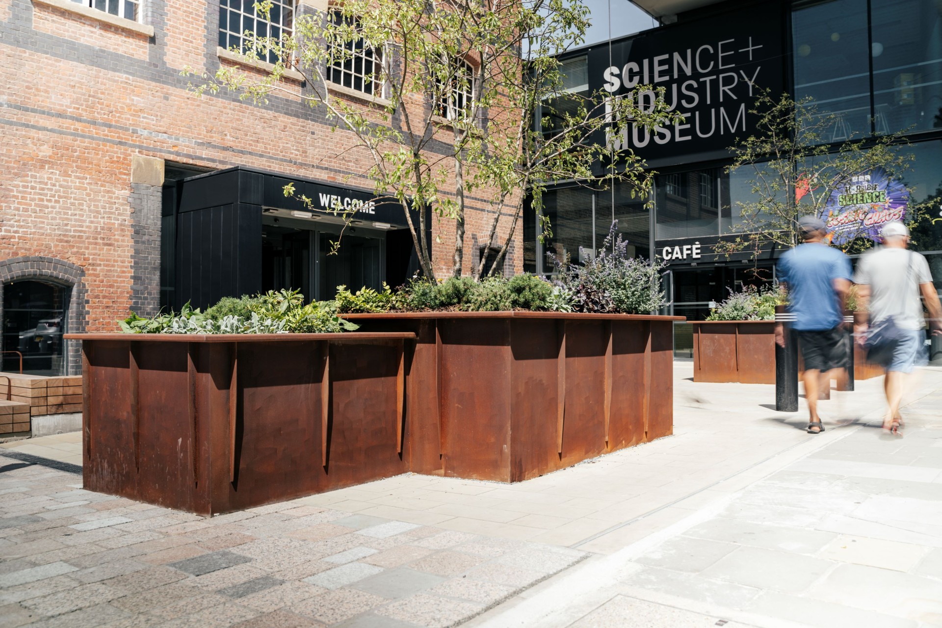 Exterior entrance of the Science and Industry Museum, featuring a historic brick building connected to a modern glass facade with large "Science + Industry Museum" signage. Rust-coloured metal planters with trees and greenery line the walkway, while two blurred pedestrians walk past the museum entrance on a sunny day.