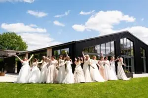 A group of brides in a variety of white and ivory wedding dresses stand laughing and posing with their arms raised on a lawn outside Shottle Hall's new modern black barn wedding venue.