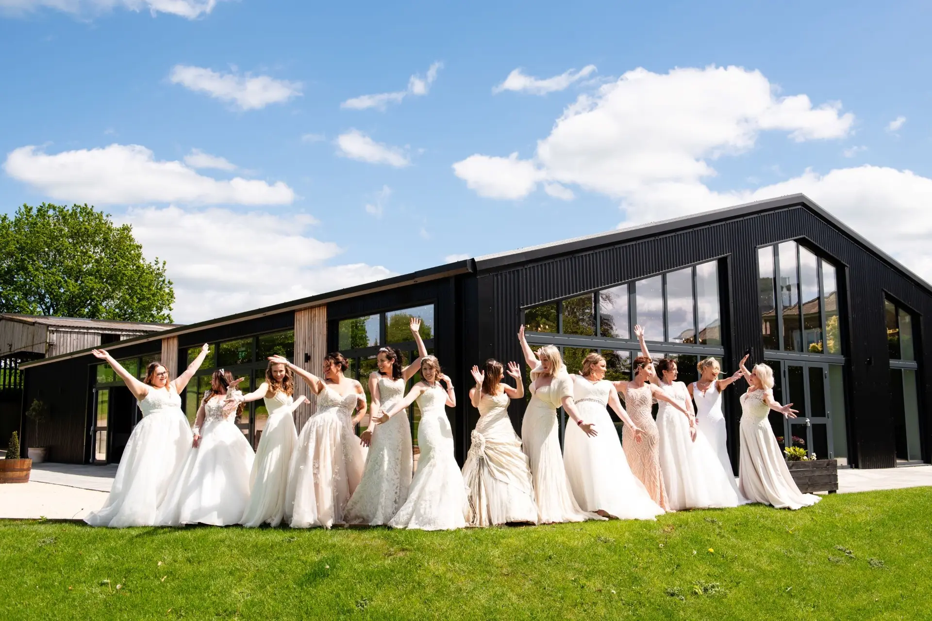 A group of brides in a variety of white and ivory wedding dresses stand laughing and posing with their arms raised on a lawn outside Shottle Hall's new modern black barn wedding venue.