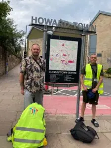 Stuart and Xavier stand with their bags beside the Glossop Town Centre map preparing to begin their 102 mile walk.