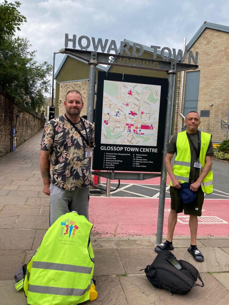 Stuart and Xavier stand with their bags beside the Glossop Town Centre map preparing to begin their 102 mile walk.
