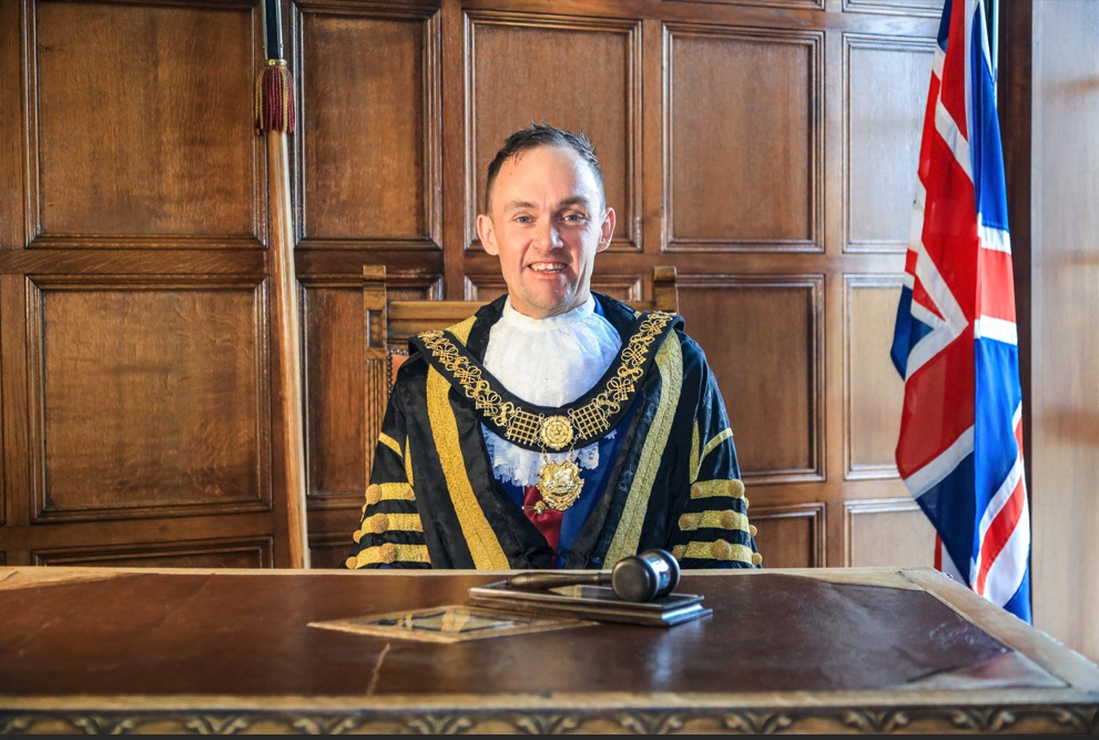 Mayor of Derby Cllr Jonathan Smale wearing ceremonial robes and chain of office, seated behind a desk with a civic gavel in the Mayor's Parlour, with a Union Jack flag in the background.