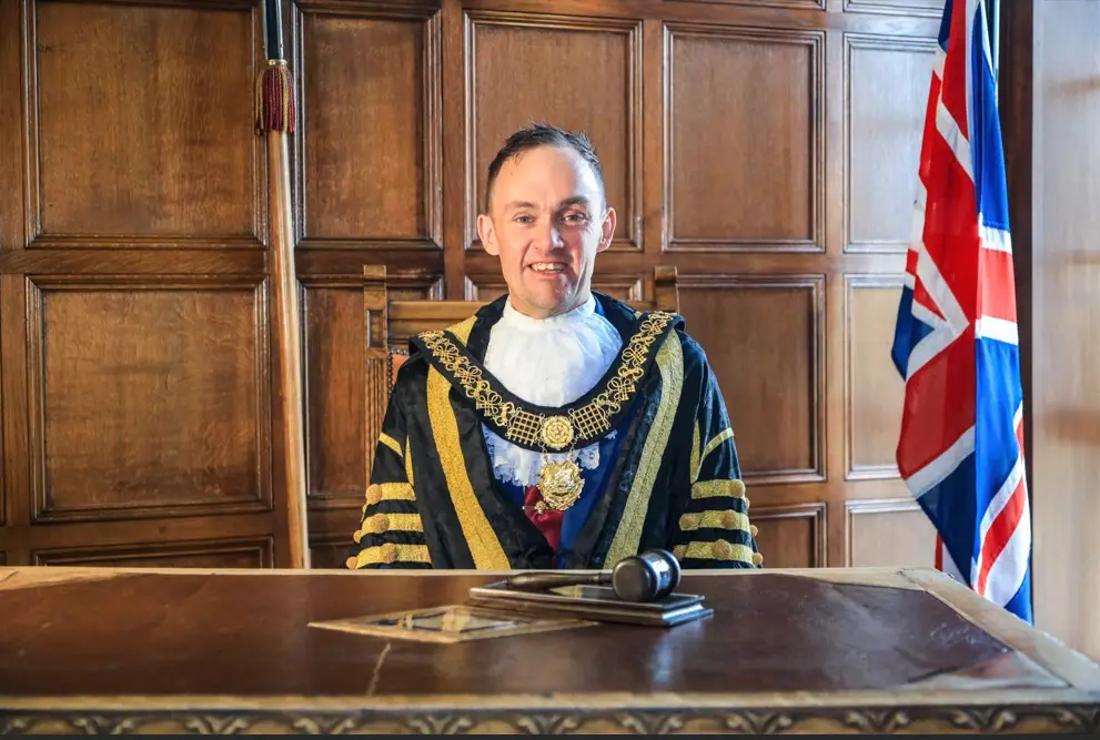 Mayor of Derby Cllr Jonathan Smale wearing ceremonial robes and chain of office, seated behind a desk with a civic gavel in the Mayor's Parlour, with a Union Jack flag in the background.