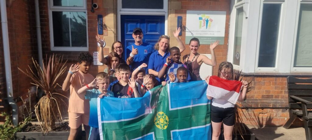Children and staff from Derbyshire Children’s Holiday Centre smile and wave with Grzegorz Pacho outside the Skegness centre while holding a large Derbyshire flag and a Polish flag.